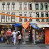 A bustling street market scene with several stalls displaying various items for sale. People of different ages are walking and exploring the market. Traditional shophouses with colorful window shutters and architectural details form the backdrop. Strings of red lanterns hang above the street, adding a festive atmosphere.
