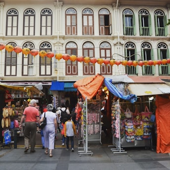 A bustling street market scene with several stalls displaying various items for sale. People of different ages are walking and exploring the market. Traditional shophouses with colorful window shutters and architectural details form the backdrop. Strings of red lanterns hang above the street, adding a festive atmosphere.