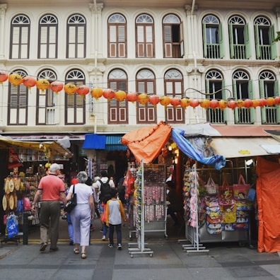 A bustling street market scene with several stalls displaying various items for sale. People of different ages are walking and exploring the market. Traditional shophouses with colorful window shutters and architectural details form the backdrop. Strings of red lanterns hang above the street, adding a festive atmosphere.