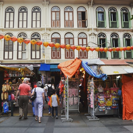 A colorful street scene in Geylang Serai with community members chatting and smiling around market stalls.