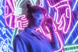 A person wearing futuristic glasses stands in front of vibrant neon lights. The background features bold electric purple, blue, and pink colors with abstract designs, possibly depicting elements like rockets or space themes. The individual is dressed in a casual outfit with a shirt and sweater, and their expression appears contemplative.