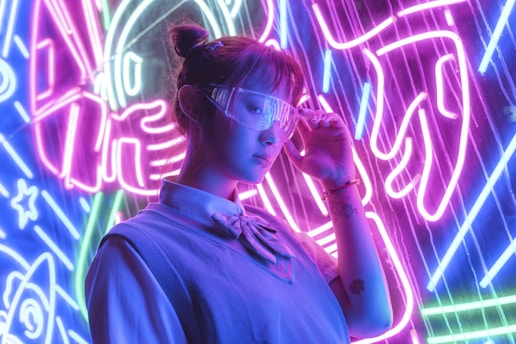 A person wearing futuristic glasses stands in front of vibrant neon lights. The background features bold electric purple, blue, and pink colors with abstract designs, possibly depicting elements like rockets or space themes. The individual is dressed in a casual outfit with a shirt and sweater, and their expression appears contemplative.