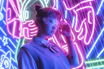 A person wearing futuristic glasses stands in front of vibrant neon lights. The background features bold electric purple, blue, and pink colors with abstract designs, possibly depicting elements like rockets or space themes. The individual is dressed in a casual outfit with a shirt and sweater, and their expression appears contemplative.
