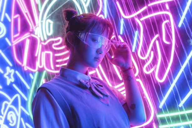 A person wearing futuristic glasses stands in front of vibrant neon lights. The background features bold electric purple, blue, and pink colors with abstract designs, possibly depicting elements like rockets or space themes. The individual is dressed in a casual outfit with a shirt and sweater, and their expression appears contemplative.