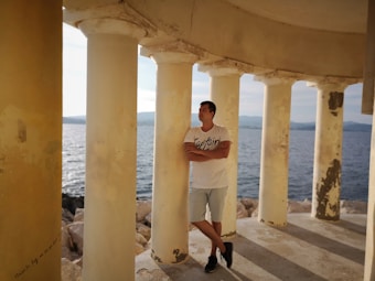 A person is standing with arms crossed, leaning against a column in a structure with multiple pillars. The background features a sea view under a clear sky, creating a serene and contemplative atmosphere.
