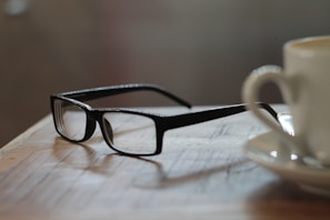 A pair of black eyeglasses rests on a wooden surface, next to a white ceramic cup with a saucer, likely containing coffee. The scene is in soft focus, giving a cozy and relaxed atmosphere. The eyewear and cup are positioned in close proximity, hinting at a casual or contemplative setting like a coffee shop or home.