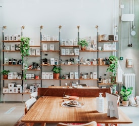 A cozy interior space featuring a wooden shelf filled with various coffee-related products and potted plants. The shelves are stocked with coffee bags, brewing devices, and decor items, all organized neatly. In front of the shelves, a wooden table is laid out with a few items, including a hand sanitizer bottle, a plate with a dessert, and a box.