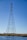 A tall metal transmission tower standing in a body of water with clear blue sky in the background. The structure has a lattice design and supports several high-voltage power lines. The surrounding area includes water and grassy vegetation.