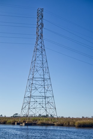 A tall metal transmission tower standing in a body of water with clear blue sky in the background. The structure has a lattice design and supports several high-voltage power lines. The surrounding area includes water and grassy vegetation.