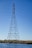 A tall metal transmission tower standing in a body of water with clear blue sky in the background. The structure has a lattice design and supports several high-voltage power lines. The surrounding area includes water and grassy vegetation.