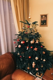 A decorated Christmas tree stands in a living room corner, adorned with various ornaments such as red and gold baubles, a star, and festive figurines. The room features warm tones with a brown leather sofa, beige curtains, and a floral painting on the wall.