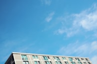 A modern apartment building is visible against a bright blue sky with wispy clouds. The building has multiple windows, and the facade consists of light-colored panels.