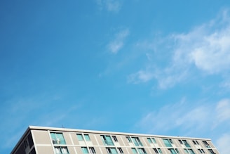 A modern apartment building is visible against a bright blue sky with wispy clouds. The building has multiple windows, and the facade consists of light-colored panels.