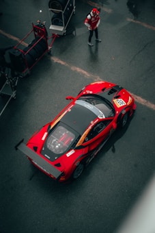 A bright red sports car is parked on a dark asphalt surface in a pit lane, with a worker in a red jacket and cap standing nearby. There are spare tires stacked on a trolley next to the car, indicating a racing environment.