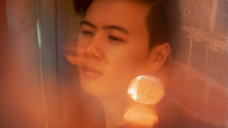 A person with a contemplative expression is captured in a soft-focus setting, with warm tones and reflections surrounding them. The scene is partially blurred, giving it a dreamy quality, with light bokeh effects contributing to the ethereal atmosphere.