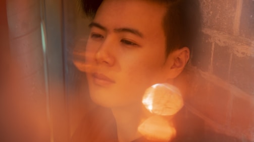A person with a contemplative expression is captured in a soft-focus setting, with warm tones and reflections surrounding them. The scene is partially blurred, giving it a dreamy quality, with light bokeh effects contributing to the ethereal atmosphere.