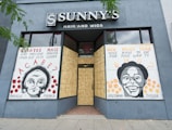 The storefront of a hair and wigs shop features two large murals with portraits of Marielle Franco and Breonna Taylor. The artwork includes text in both Portuguese and English, posing poignant questions about social justice and the impact of violence. The shop windows are boarded up, and the murals are displayed against a gray-blue exterior wall.