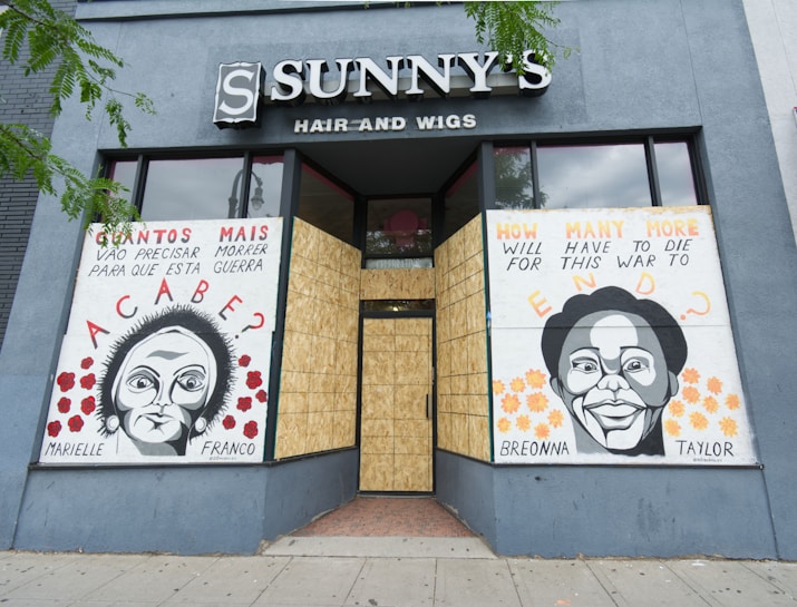 The storefront of a hair and wigs shop features two large murals with portraits of Marielle Franco and Breonna Taylor. The artwork includes text in both Portuguese and English, posing poignant questions about social justice and the impact of violence. The shop windows are boarded up, and the murals are displayed against a gray-blue exterior wall.