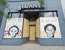 The storefront of a hair and wigs shop features two large murals with portraits of Marielle Franco and Breonna Taylor. The artwork includes text in both Portuguese and English, posing poignant questions about social justice and the impact of violence. The shop windows are boarded up, and the murals are displayed against a gray-blue exterior wall.