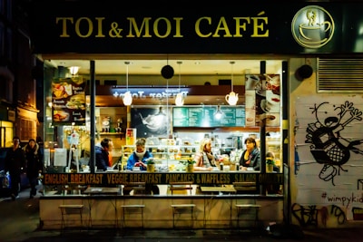 A cozy café named 'Toi & Moi Café' has a warm and inviting interior with patrons seated at tables. The café showcases an array of menu options such as English breakfast and avocado sandwiches. The exterior is lively with a large window revealing the bustling activity inside, while outside a street-style wall art adds to the urban atmosphere.