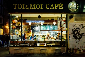 A cozy café named 'Toi & Moi Café' has a warm and inviting interior with patrons seated at tables. The café showcases an array of menu options such as English breakfast and avocado sandwiches. The exterior is lively with a large window revealing the bustling activity inside, while outside a street-style wall art adds to the urban atmosphere.