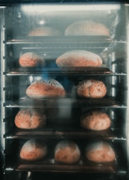 Multiple loaves of bread are baking inside an industrial oven, arranged on metal racks. The bread has a golden-brown crust and appears to be in various shapes and sizes.
