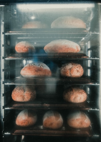 Multiple loaves of bread are baking inside an industrial oven, arranged on metal racks. The bread has a golden-brown crust and appears to be in various shapes and sizes.