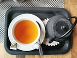 A cup of tea and a black teapot sit on a black tray with a black and white checkered background. The tea is in a white cup and is accompanied by a teaspoon and a packet of sugar. The teapot has a textured, dotted pattern and a curved handle.