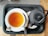A cup of tea and a black teapot sit on a black tray with a black and white checkered background. The tea is in a white cup and is accompanied by a teaspoon and a packet of sugar. The teapot has a textured, dotted pattern and a curved handle.