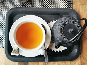 A cup of tea and a black teapot sit on a black tray with a black and white checkered background. The tea is in a white cup and is accompanied by a teaspoon and a packet of sugar. The teapot has a textured, dotted pattern and a curved handle.