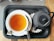 A cup of tea and a black teapot sit on a black tray with a black and white checkered background. The tea is in a white cup and is accompanied by a teaspoon and a packet of sugar. The teapot has a textured, dotted pattern and a curved handle.