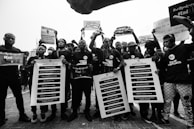 A group of people are gathered, holding protest signs and banners demanding social and political change. They are wearing coordinated clothing that suggests unity in their cause, with messages related to ending violence and reforms. The setting appears to be outdoors, possibly during a protest march.
