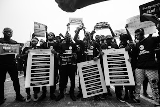 A group of people are gathered, holding protest signs and banners demanding social and political change. They are wearing coordinated clothing that suggests unity in their cause, with messages related to ending violence and reforms. The setting appears to be outdoors, possibly during a protest march.