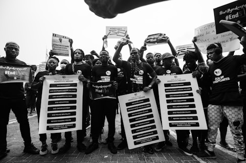 A group of people are gathered, holding protest signs and banners demanding social and political change. They are wearing coordinated clothing that suggests unity in their cause, with messages related to ending violence and reforms. The setting appears to be outdoors, possibly during a protest march.