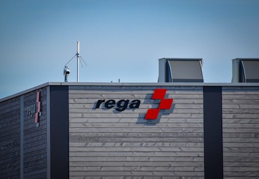 A modern building facade made of horizontal wooden planks features the word 'rega' accompanied by a red geometric symbol. The structure is equipped with a camera and an antenna on the roof, and has large, metallic-looking air vents.