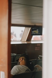 A person wearing a mask and cap is seated inside a transportation vehicle, possibly a ferry or bus. The interior is dimly lit with rows of green seats and wood paneled walls. A television screen displays a person wearing a yellow shirt. Through the window, blurred silhouettes of people are visible against a bright background.