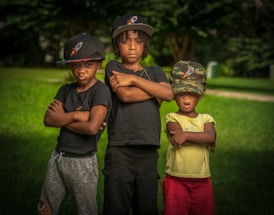 Three children stand outdoors on a grassy field, each with their arms crossed. They are wearing hats featuring a logo with a rocket. The child on the left and the one in the middle wear black shirts, while the child on the right is in a yellow shirt and red shorts. The background shows trees with a blurred effect, giving a natural setting.