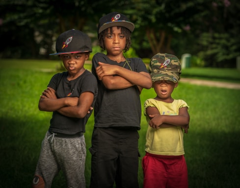 Three children stand outdoors on a grassy field, each with their arms crossed. They are wearing hats featuring a logo with a rocket. The child on the left and the one in the middle wear black shirts, while the child on the right is in a yellow shirt and red shorts. The background shows trees with a blurred effect, giving a natural setting.