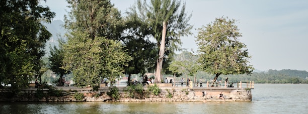 A lakeside scene features a row of lush green trees on a stone embankment, with people sitting and enjoying the view. The background includes distant hills under a clear sky, creating a serene and peaceful atmosphere.