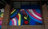 A colorful mural featuring a stylized raised fist in green and black with a red heart in the palm. Surrounding the fist are vibrant rainbow stripes and a trans pride flag. The words 'Stand Together' are written above in elegant script.
