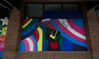 A colorful mural featuring a stylized raised fist in green and black with a red heart in the palm. Surrounding the fist are vibrant rainbow stripes and a trans pride flag. The words 'Stand Together' are written above in elegant script.