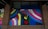 A colorful mural featuring a stylized raised fist in green and black with a red heart in the palm. Surrounding the fist are vibrant rainbow stripes and a trans pride flag. The words 'Stand Together' are written above in elegant script.