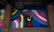 A colorful mural featuring a stylized raised fist in green and black with a red heart in the palm. Surrounding the fist are vibrant rainbow stripes and a trans pride flag. The words 'Stand Together' are written above in elegant script.