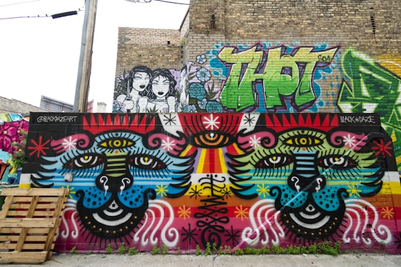 A vibrant, colorful mural is painted on a brick wall in an urban setting, featuring large, symmetric faces with multiple eyes, surrounded by abstract patterns and vivid details. Above the primary artwork, there is additional graffiti including large, stylized lettering and a sketch of two cartoon-like figures. A wooden pallet leans against the wall in the foreground.