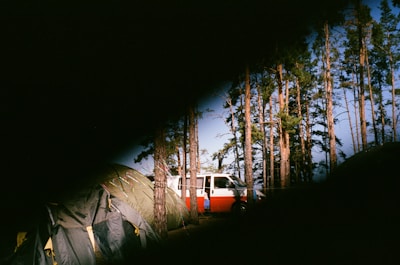 A camping scene with a tent and a red and white van surrounded by tall pine trees. The image has a partial vignette effect, giving it a mysterious and obscured appearance.