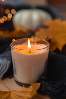 A close-up of a warm butterscotch candle glowing softly on a rustic wooden table with autumn leaves scattered around.