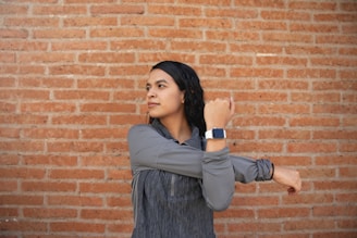 A person is stretching their arm across their chest, wearing a gray jacket and a smartwatch. The background consists of a textured brick wall, creating a casual urban setting.