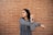 A person is stretching their arm across their chest, wearing a gray jacket and a smartwatch. The background consists of a textured brick wall, creating a casual urban setting.