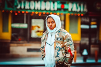 A person wearing a camouflage jacket and a grey hoodie stands on a city street. The background includes a sign for Stromboli and Pasta, with bright lights and a blurred effect, indicating an urban setting.