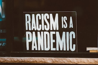 A sign with bold, white text reading 'RACISM IS A PANDEMIC' placed against a dark background. The lettering is highly visible and occupies most of the image. A small part of a book is visible in the bottom right corner.